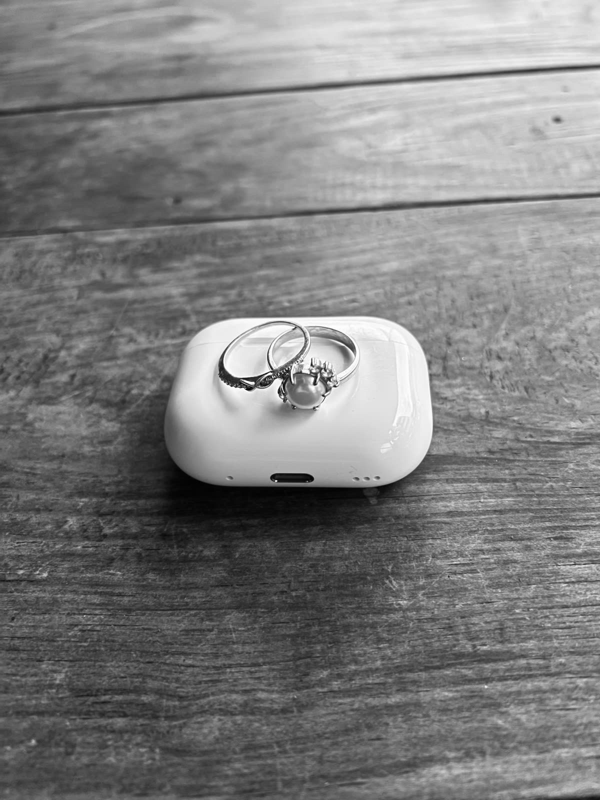 Two rings placed on top of a white wireless earbud case, set on a wooden surface in black and white. — 에어팟 5 국내 출시, 한국 예상가는 이렇게 계산합니다